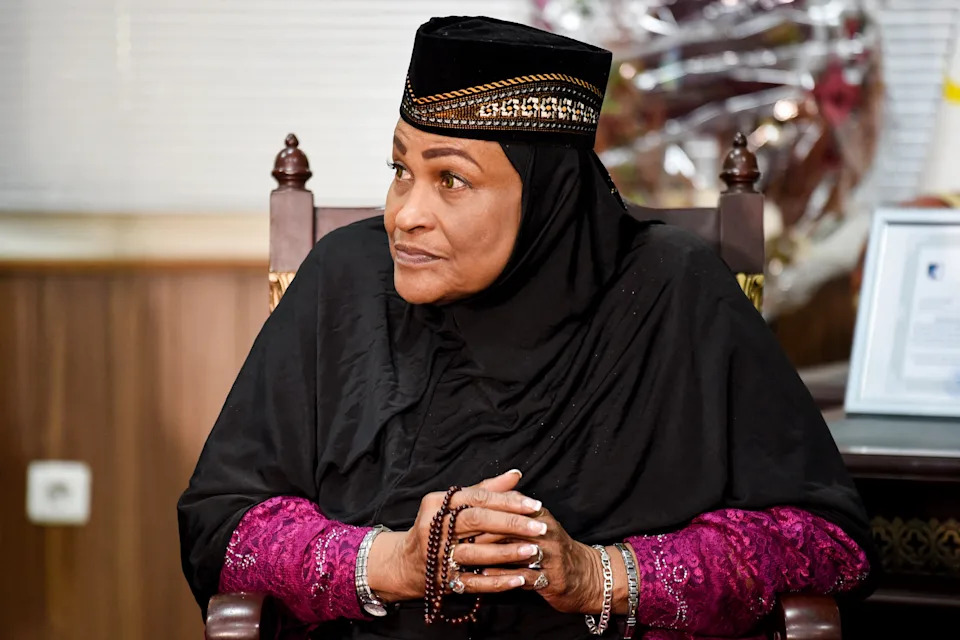 Khalilah Ali seated, wearing traditional attire with a beaded headpiece, holding prayer beads, looking thoughtfully to the side
