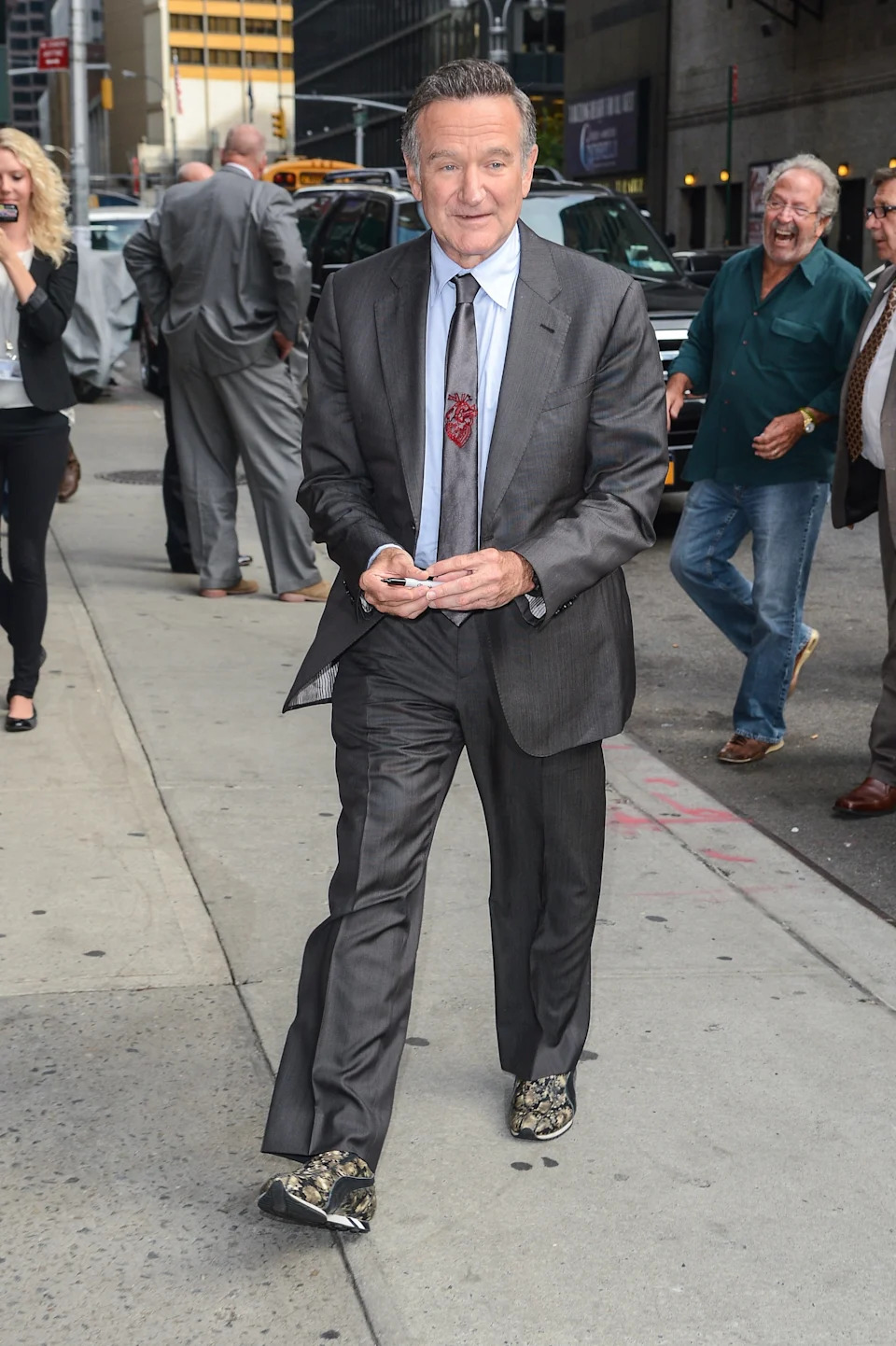 Robin Williams in a suit with patterned shoes walking on city sidewalk, smiling slightly, surrounded by pedestrian activity