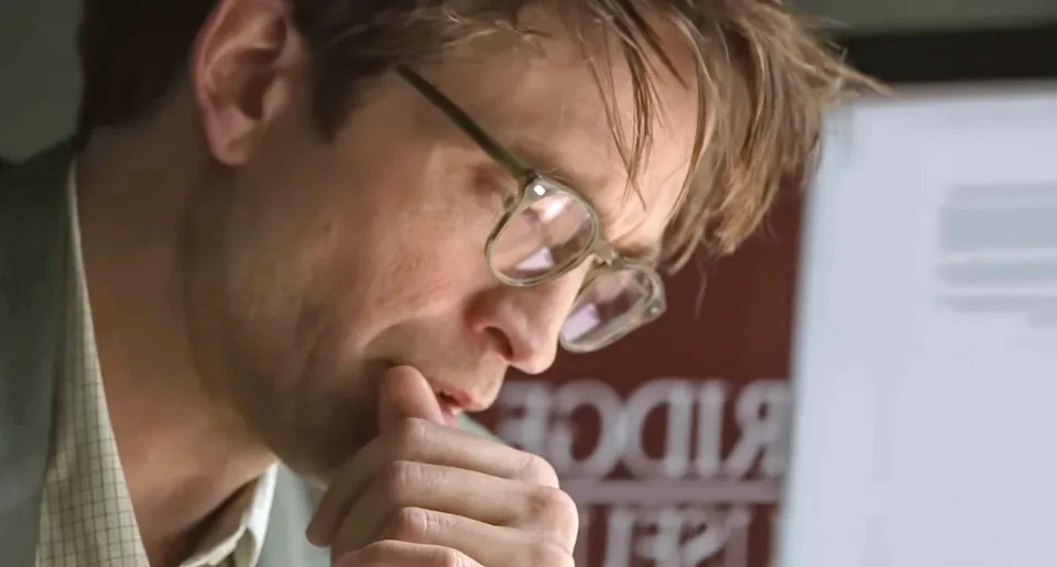 Person with glasses looks thoughtful, resting chin on hand, in front of a computer screen