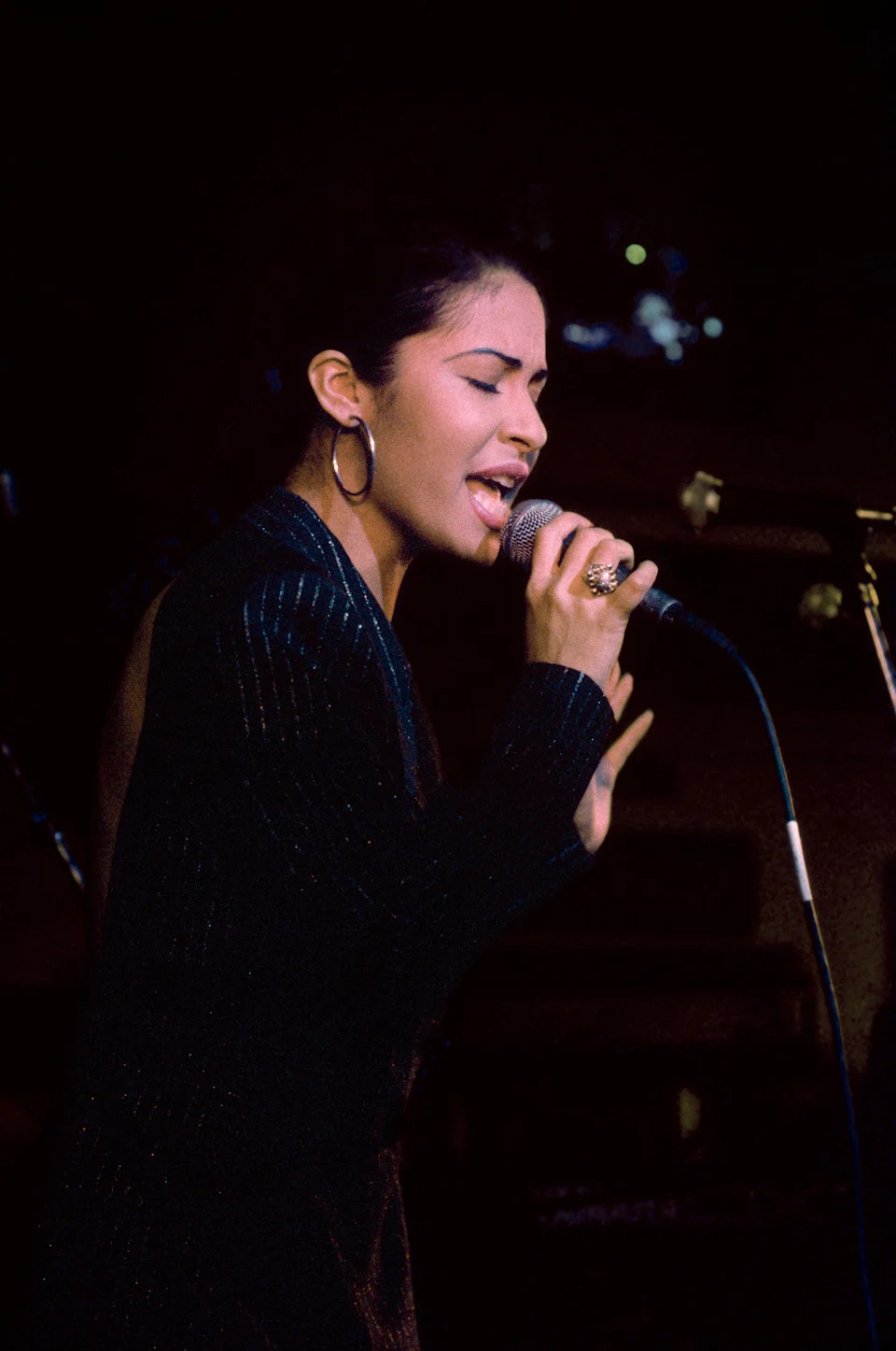 Selena performing passionately with a microphone, wearing a textured, elegant outfit and large hoop earrings