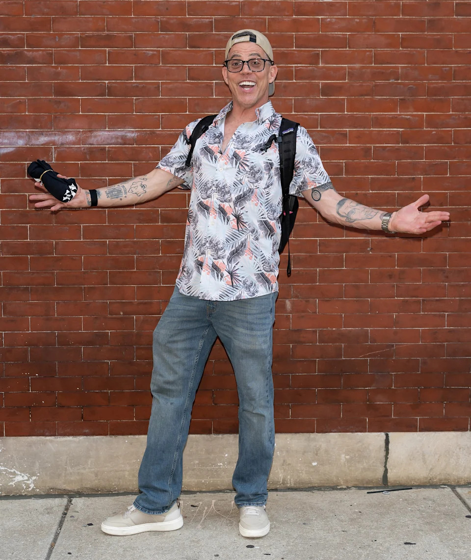 Person posing happily against a brick wall, wearing a floral shirt, jeans, cap, and glasses, with a backpack and holding a small device