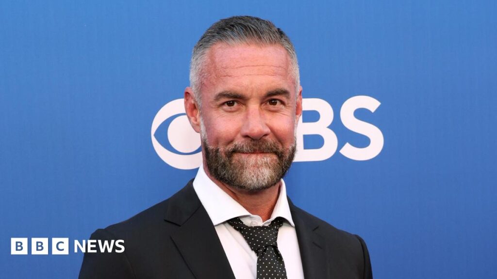 Police stop scam victim selling home to ‘wed’ actor Jay Harrington Actor Jay Harrington wearing a black suit with a white shirt and dark patterned tie. He is standing in front of a poster board which is blue and has the CBS logo on it.