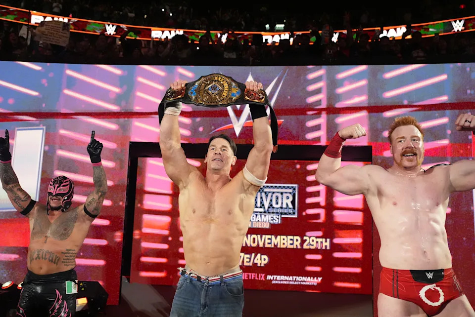 Three wrestlers celebrate in a ring; the center wrestler holds a championship belt overhead, flanked by two flexing wrestlers