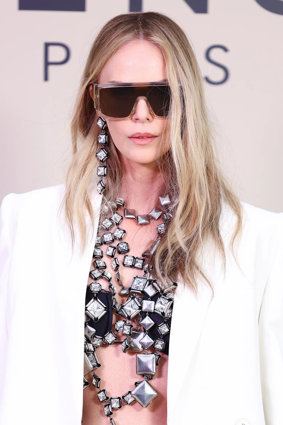 Person wearing oversized sunglasses, bold geometric jewelry, and a stylish white jacket at an event, conveying a chic, modern look