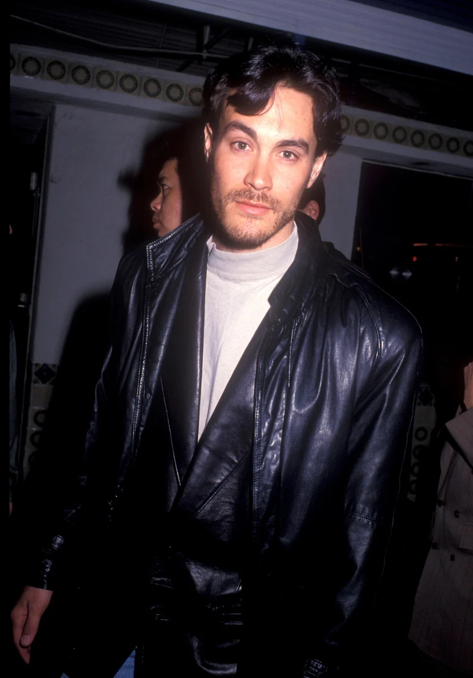 A person in a leather jacket and light turtleneck poses at a nighttime event against a dimly lit background