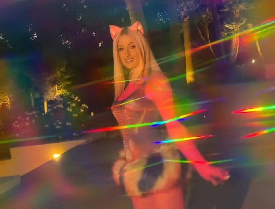 Paris Hilton wearing a playful outfit with cat ears, a corset, and a furry skirt in a vibrant, rainbow-hued setting