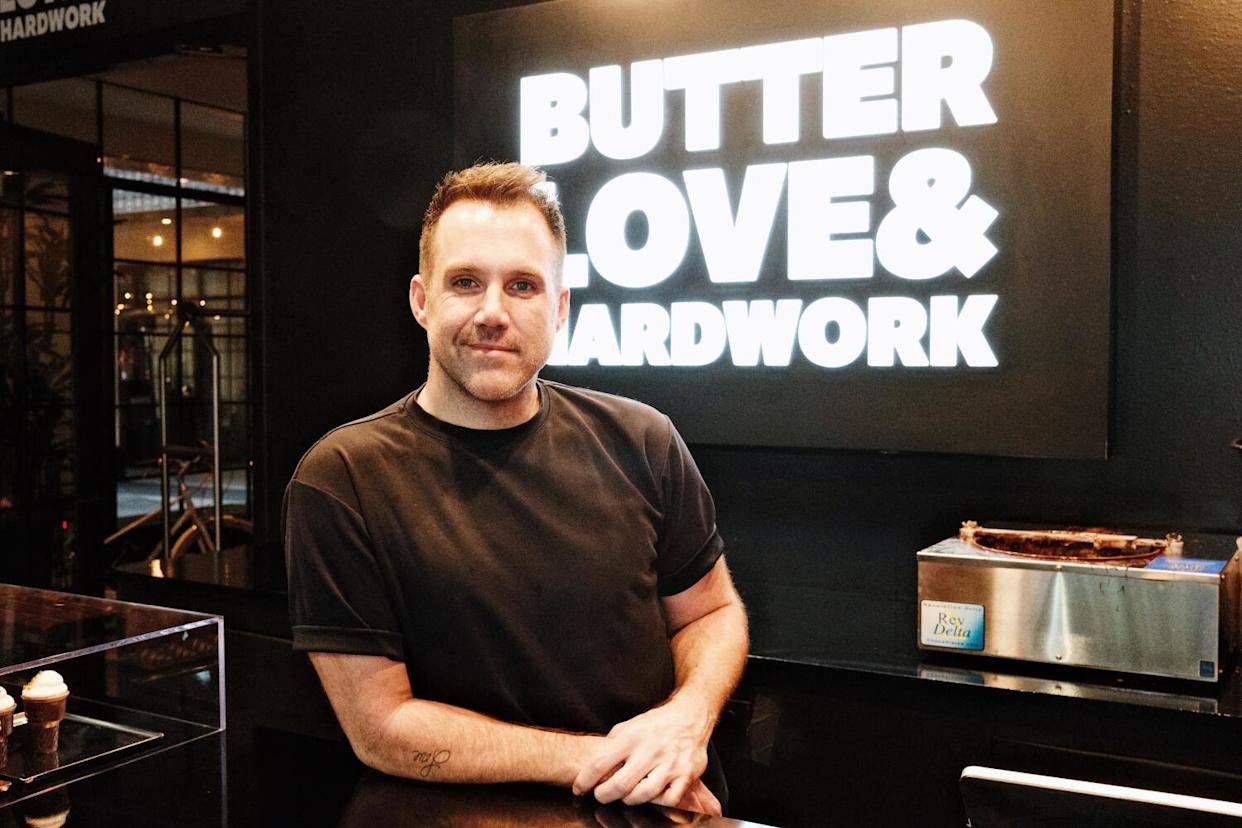 A man stands behind a reflective black counter in front of a sign that reads "BUTTER LOVE AND HARD WORK"