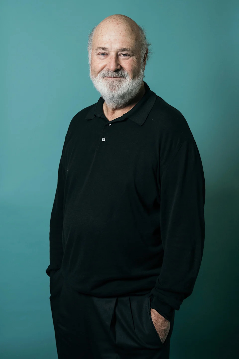 Person with a beard in a casual black shirt and pants poses with hands in pockets against a plain backdrop