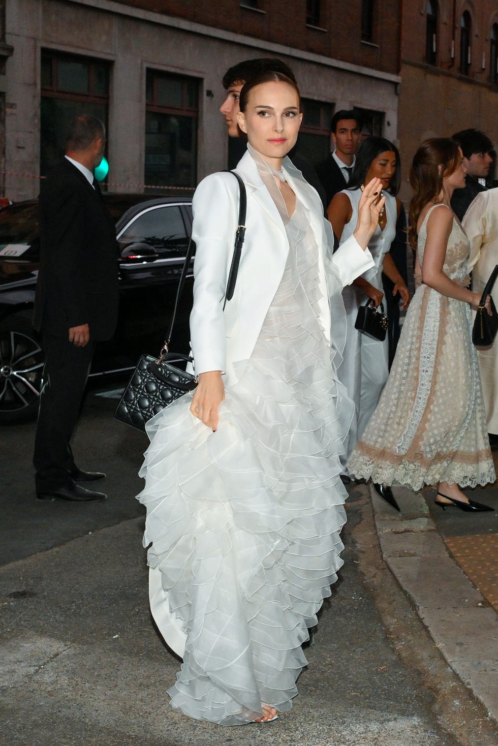 A person wearing a white layered dress and a blazer, holding a black handbag, walking on the street.