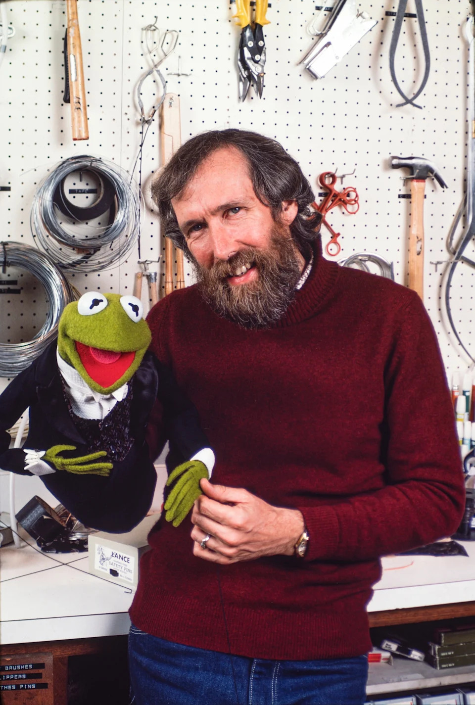 A man with a beard stands next to a puppet resembling a frog, both smiling. Tools hang on a pegboard in the background