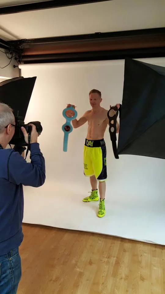 Philip Sharkey, seen from behind with a DSLR raised to his eye, photographs a shirtless boxer holding two championship belts aloft against a white studio backdrop flanked by large softboxes.