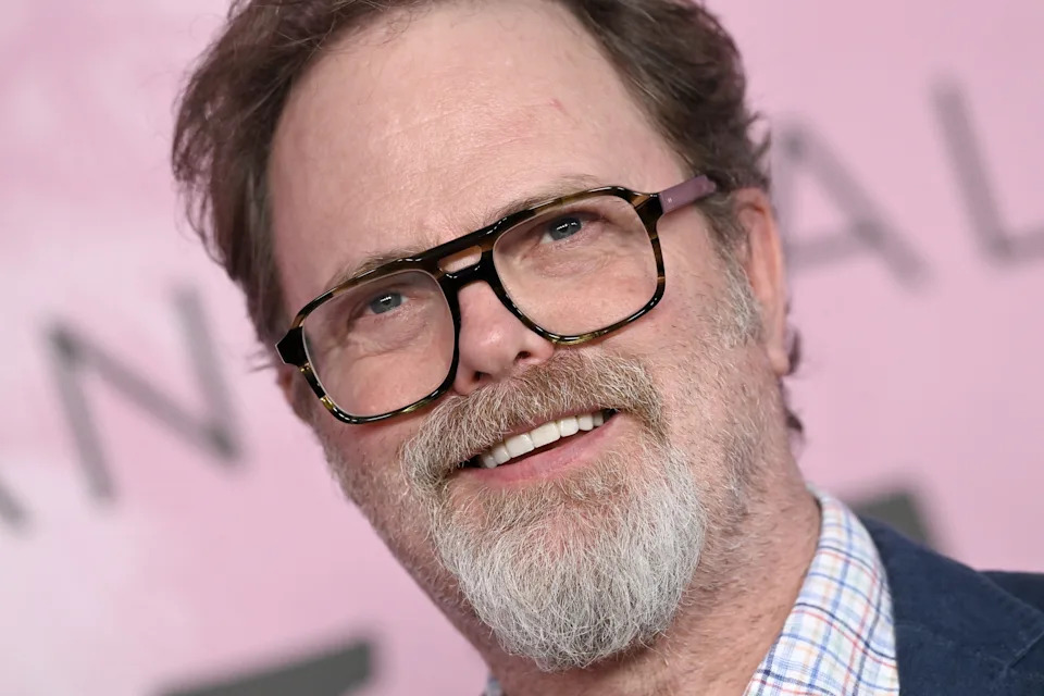 A man with glasses and a beard smiles while wearing a checked shirt and blazer at a celebrity event