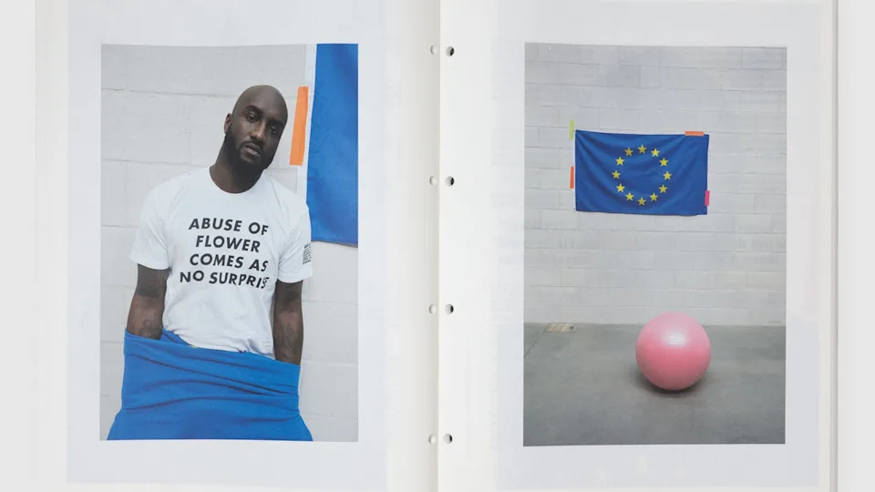 Virgil Abloh wearing a white T-shirt with text, next to a European Union flag and a pink exercise ball in a minimalist setting.