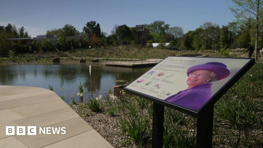 A commemorative information plaque titled "A Garden for a Queen" stands in the foreground, featuring a portrait of Queen Elizabeth II. Behind the sign is a garden landscape including a still pond, stone walkways, and a variety of trees and shrubs.