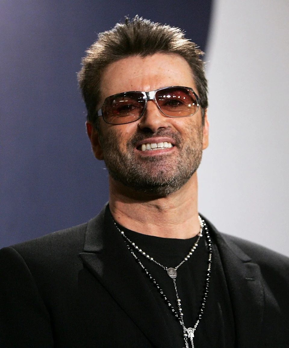 Celebrity wearing sunglasses, black suit, and layered necklaces, smiling at an event