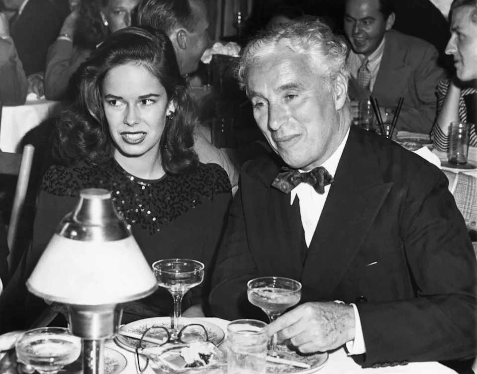 Charlie Chaplin and his wife Oona sit at a dinner table, both dressed elegantly.