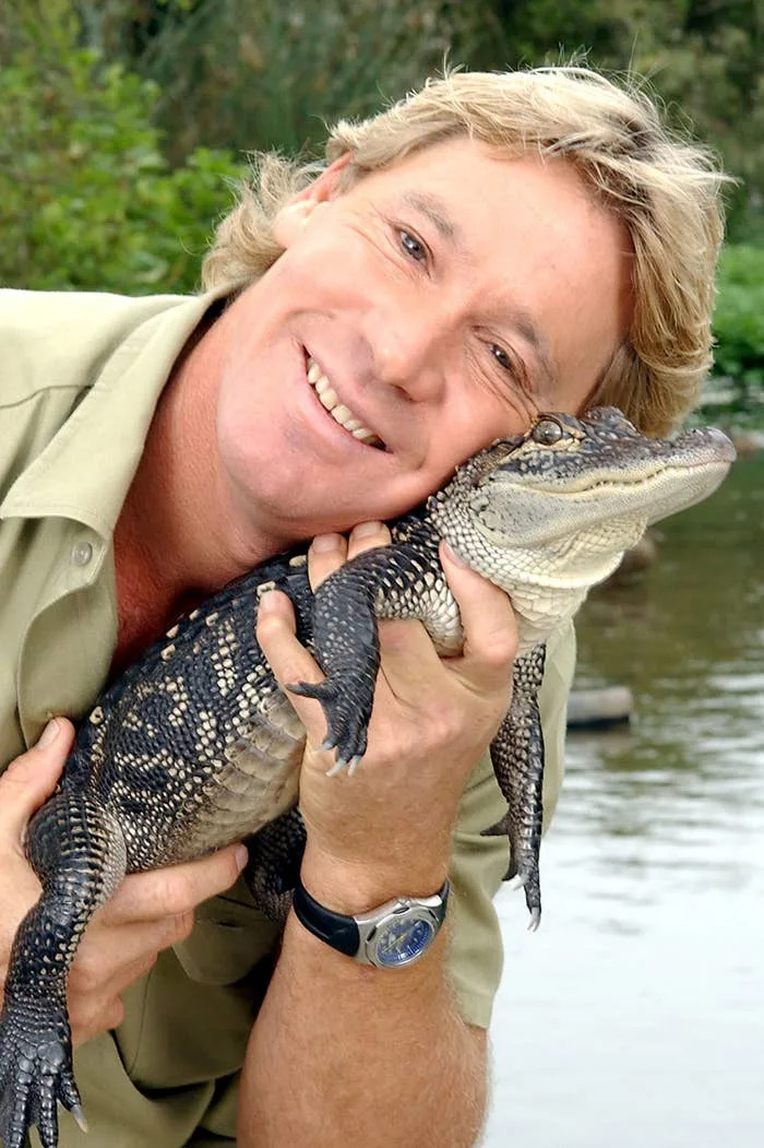 Person smiling while holding a small crocodile by a waterway. The person is wearing casual outdoor clothing