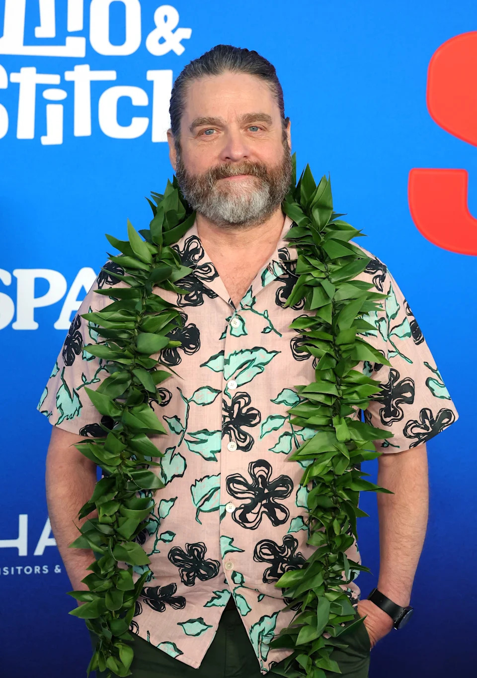 A person on a red carpet wearing a floral shirt and a leafy lei, standing in front of a blue background with text
