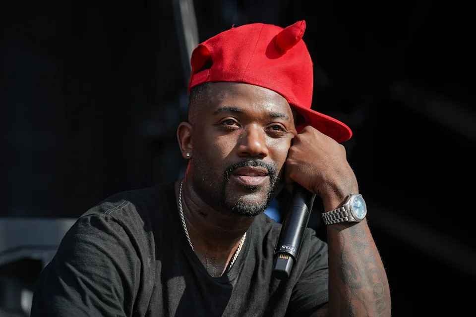 Ray J wearing a red baseball cap, tilted sideways, holding a microphone, appears thoughtful on stage