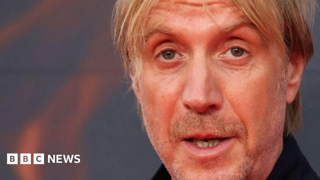 All five band members standing in side by side surrounded by yellow and orange dahlias, with more flowers behind. They all wear army green jackets and two out of five wear a cap, one a hat. Three of the men also wear sunglasses