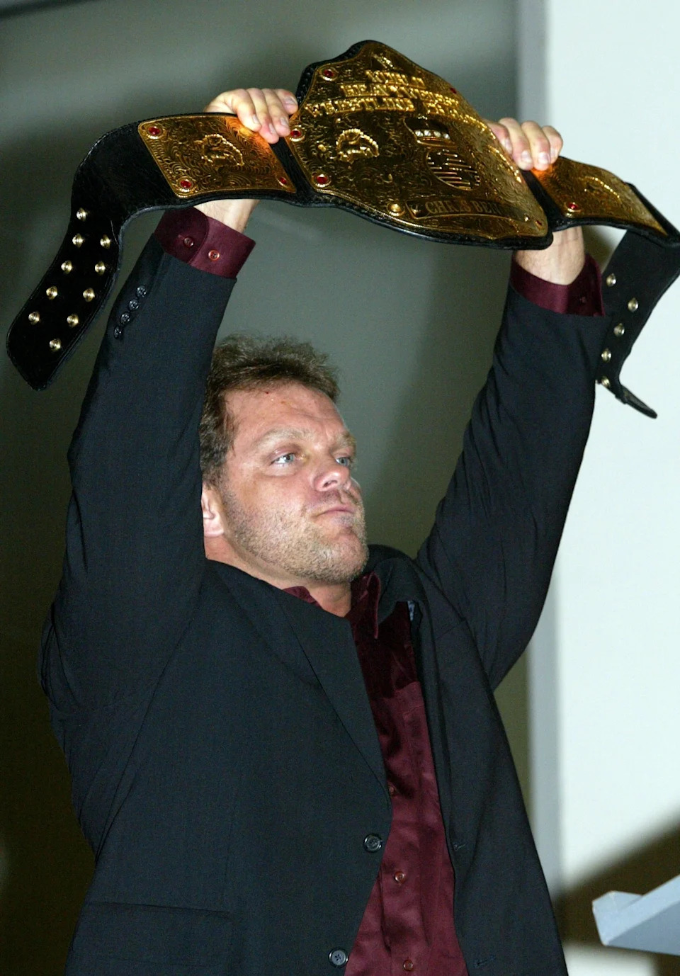Chris Benoit in a suit triumphantly holds up a large championship belt overhead