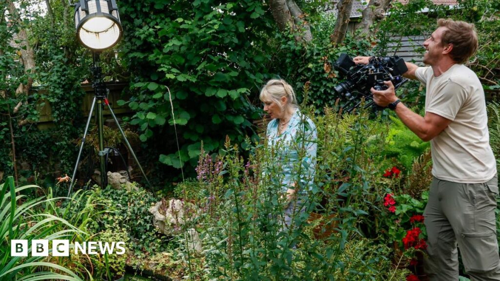 'Attenborough made a documentary about my garden' – BBC Louise Vergette wearing a blue and white blouse, with her blonde hair tied up. She is walking through her garden, which is full of different types of trees, flowers and plants.