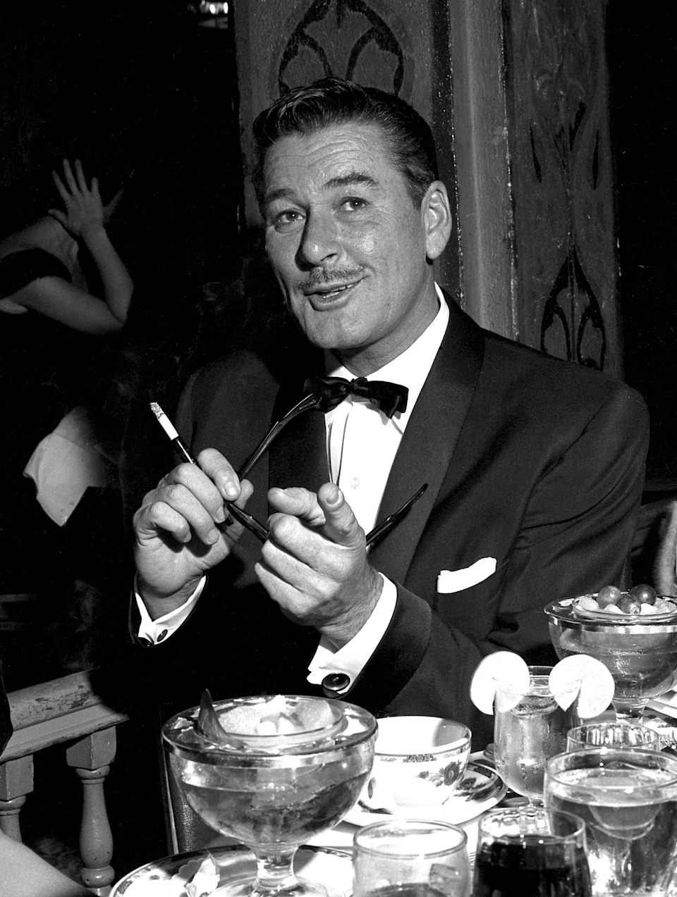 Errol Flynn in a tuxedo seated at a table with drinks and dessert, gesturing animatedly with a cigarette in hand