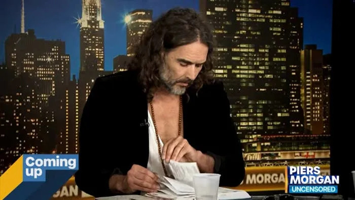 Russell Brand with long hair on a talk show set, reading a Bible with a cityscape background and show branding visible on screen