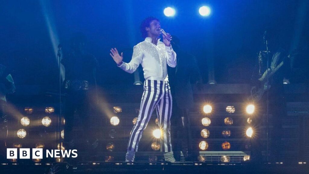 A film still showing a young man striking a pose on stage as he sings into a microphone. He has black hair in a mullet cut and wears a bright white shirt and white and blue striped trousers, which make him stand out against a dark background. The lighting is low and the shapes of his backing musicians can only just be made out. Stage lights shine in the background.