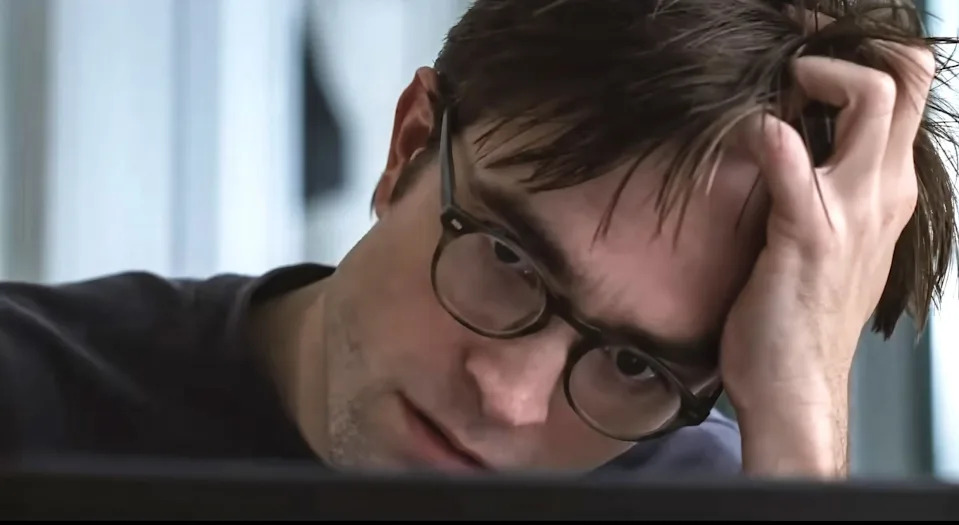 A person with glasses looks intently at a screen, resting their head on their hand, appearing thoughtful or focused