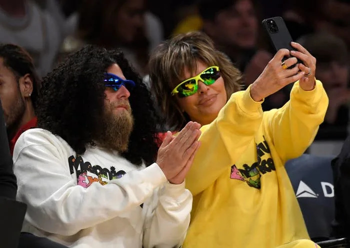 Two people wearing graphic sweatshirts and colorful sunglasses take a selfie at a public event