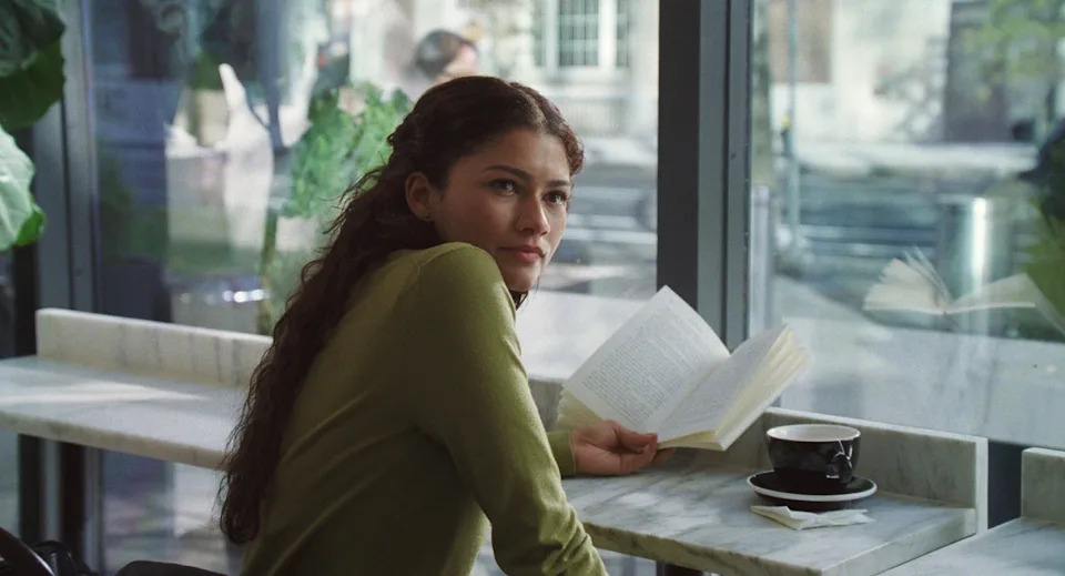 Person with long hair reading a book at a cafe, looking out the window thoughtfully. A coffee cup is on the table nearby