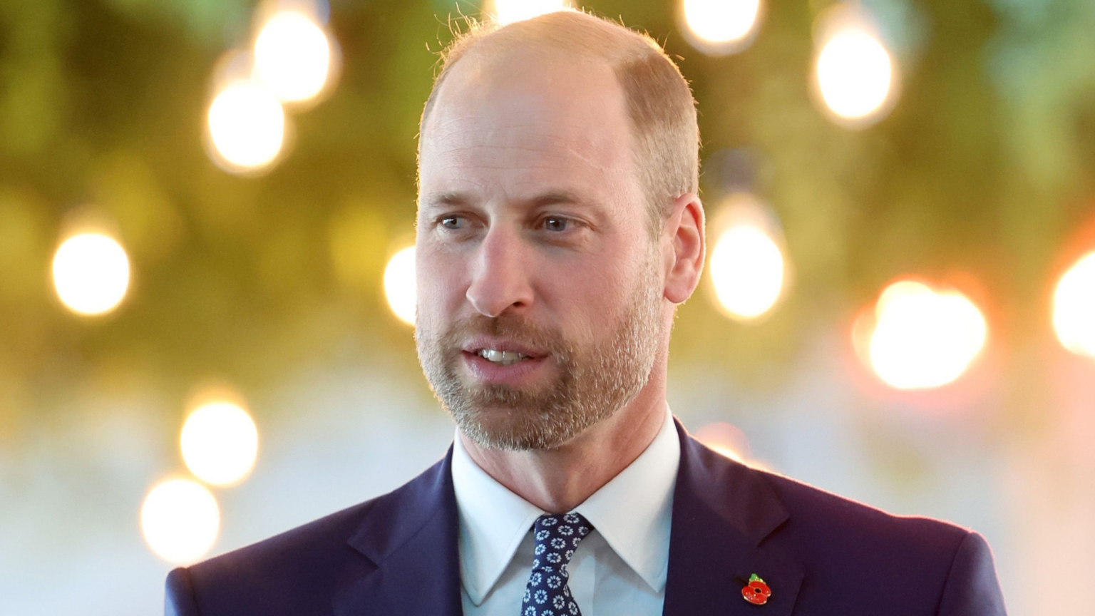 Prince William has a short beard and a bald head and wears a navy suit with a light blue shirt and a patterned tie