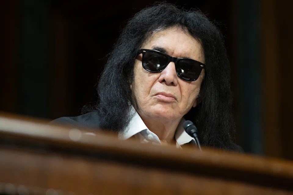 A person with long dark hair and sunglasses speaks at a microphone during a public event