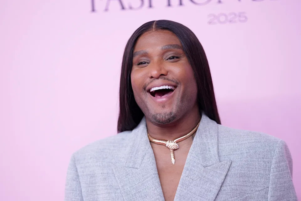 Person smiling on a red carpet, wearing a tailored, light-colored suit and a distinctive necklace