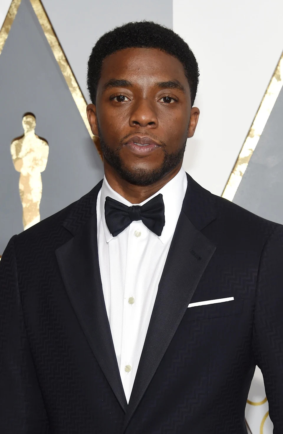 Chadwick Boseman in a tuxedo with a black bow tie poses at an event with a gold statuette backdrop