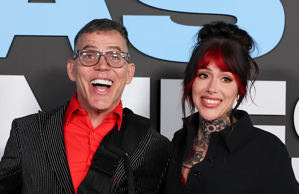 Two people posing on a red carpet. The man wears a pinstripe suit with a red shirt, and the woman has tattoos and is dressed in a dark outfit