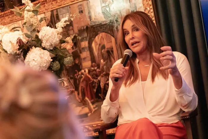 Caitlyn Jenner speaking into a microphone, wearing a blouse and skirt, with a floral arrangement and painting in the background