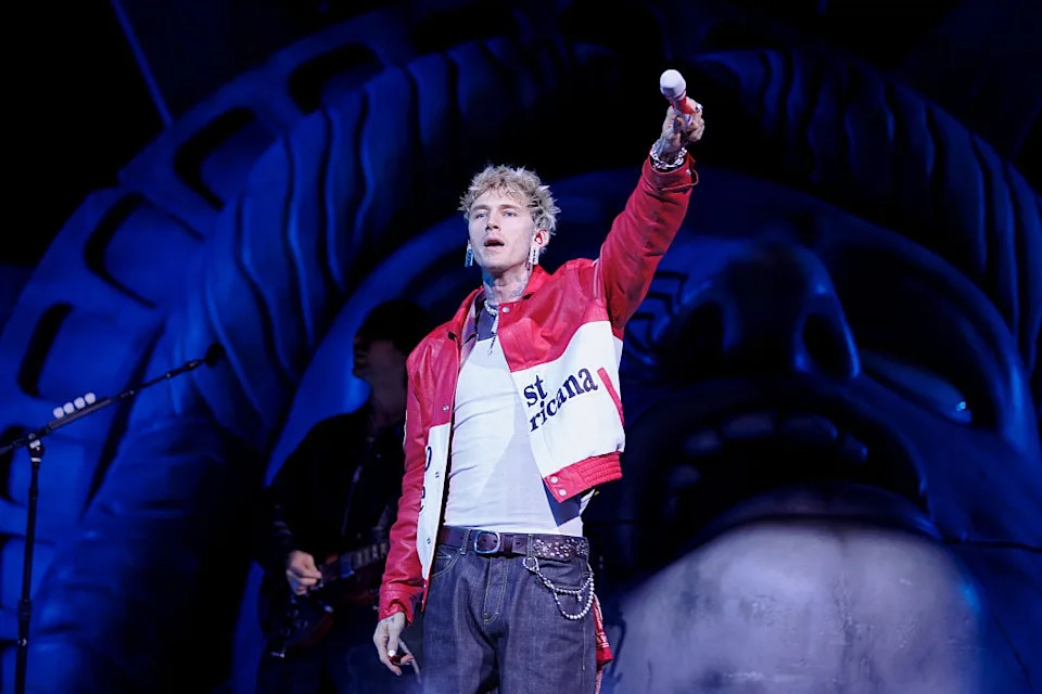 Performer on stage, wearing a stylish red and white jacket, holding a microphone with the Statue of Liberty backdrop