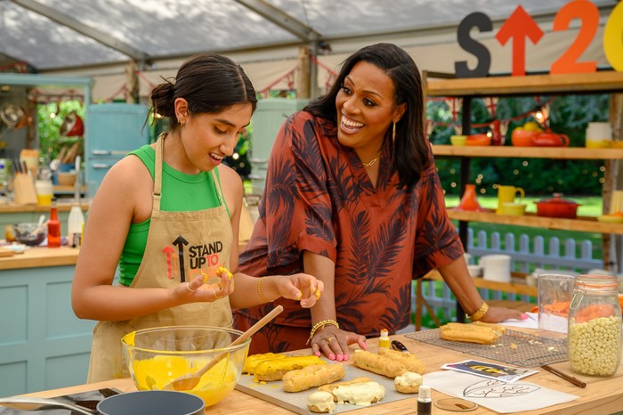 the-great-celebrity-bake-off-2026 Ambika Mod smiling down at a work service whilst she bakes whilst Alison Hammond leans on the work surface and smiles at Ambika.
