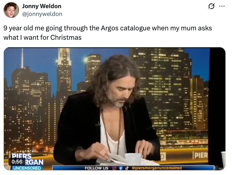 Person with long hair reads, wearing an open-collar shirt on a talk show set with a city skyline backdrop. Text above is humorous about holiday wishes