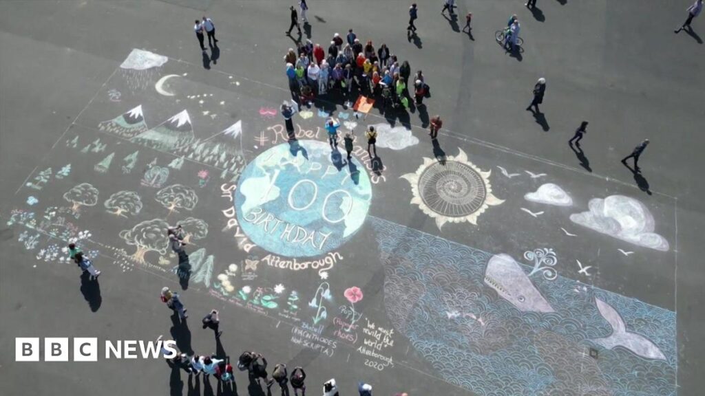 An ariel view of the artwork on Plymouth Hoe. There is Earth at the centre, with 'Happy 100 Birthday Sir David Attenborough' on it. There is a whale to Earth's right and trees and mountains to its left. A choir is gathered at the top the artwork, with people lined up taking photos on the other side.