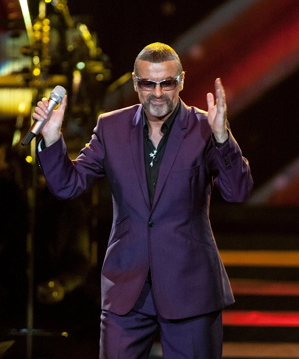 George Michael in sunglasses wearing a purple suit, holding a microphone, performing on stage