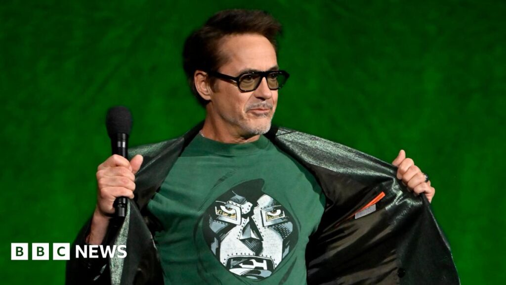 Robert Downey Jr on stage at CinemaCon, opening his jacket to reveal a T-shirt showing the masked face of his character Doctor Doom