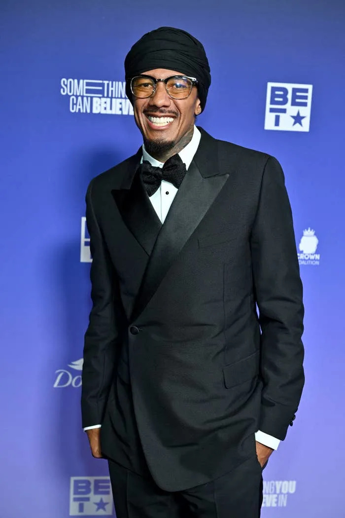 Nick Cannon on a red carpet wearing a stylish black tuxedo, white shirt, black bow tie, black turban, and glasses, smiling for photos