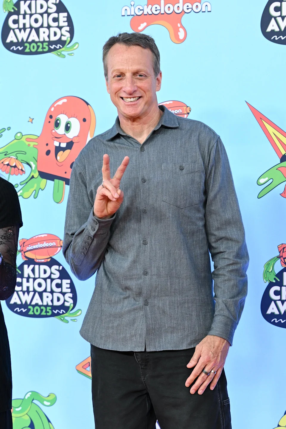 Person at Kids' Choice Awards 2025, posing with a peace sign, wearing a casual button-up shirt