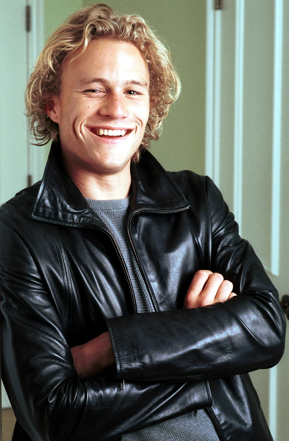 A person with wavy blonde hair smiles and poses with arms crossed, wearing a leather jacket and a light shirt