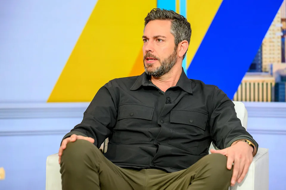 Levi sitting on a chair during a talk show interview, wearing a black shirt and olive pants, with colorful background elements behind