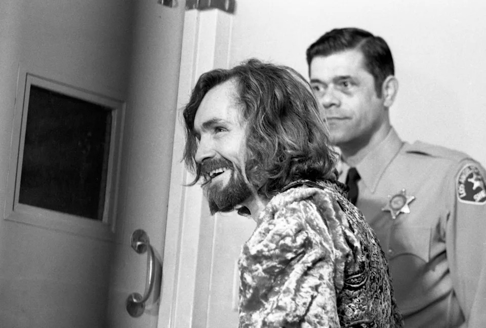 Charles Manson with long hair and beard smiling, accompanied by a uniformed officer in a hallway