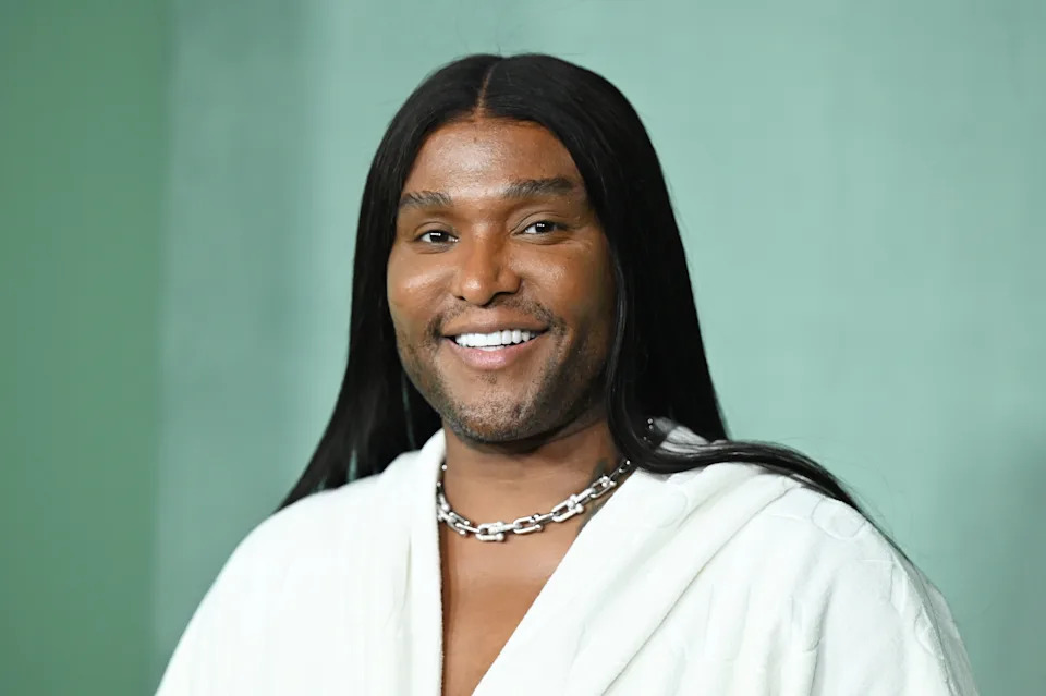 Person smiling, wearing a plush, robe-like outfit and a chunky silver chain necklace, posing against a plain backdrop